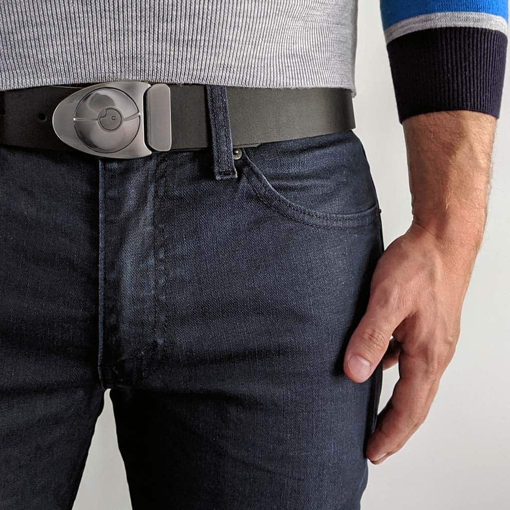 Person wearing a modern silver belt buckle shaped like an arrowhead. The black leather belt is worn with dark blue jeans and a grey sweater.